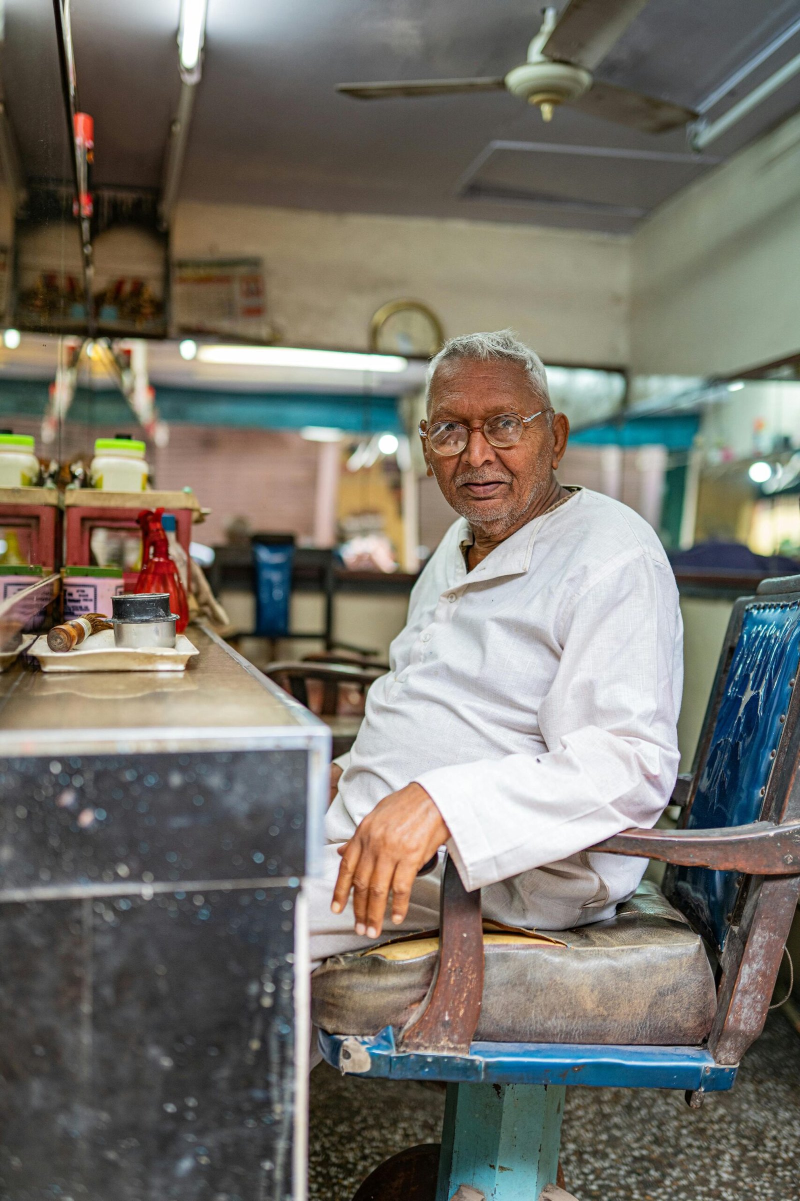Senior man in a white shirt seated in a classic Varanasi barbershop, India.